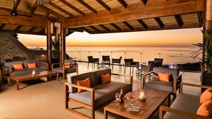 Un espacio al aire libre con mobiliario de madera, vista al océano y un atardecer.