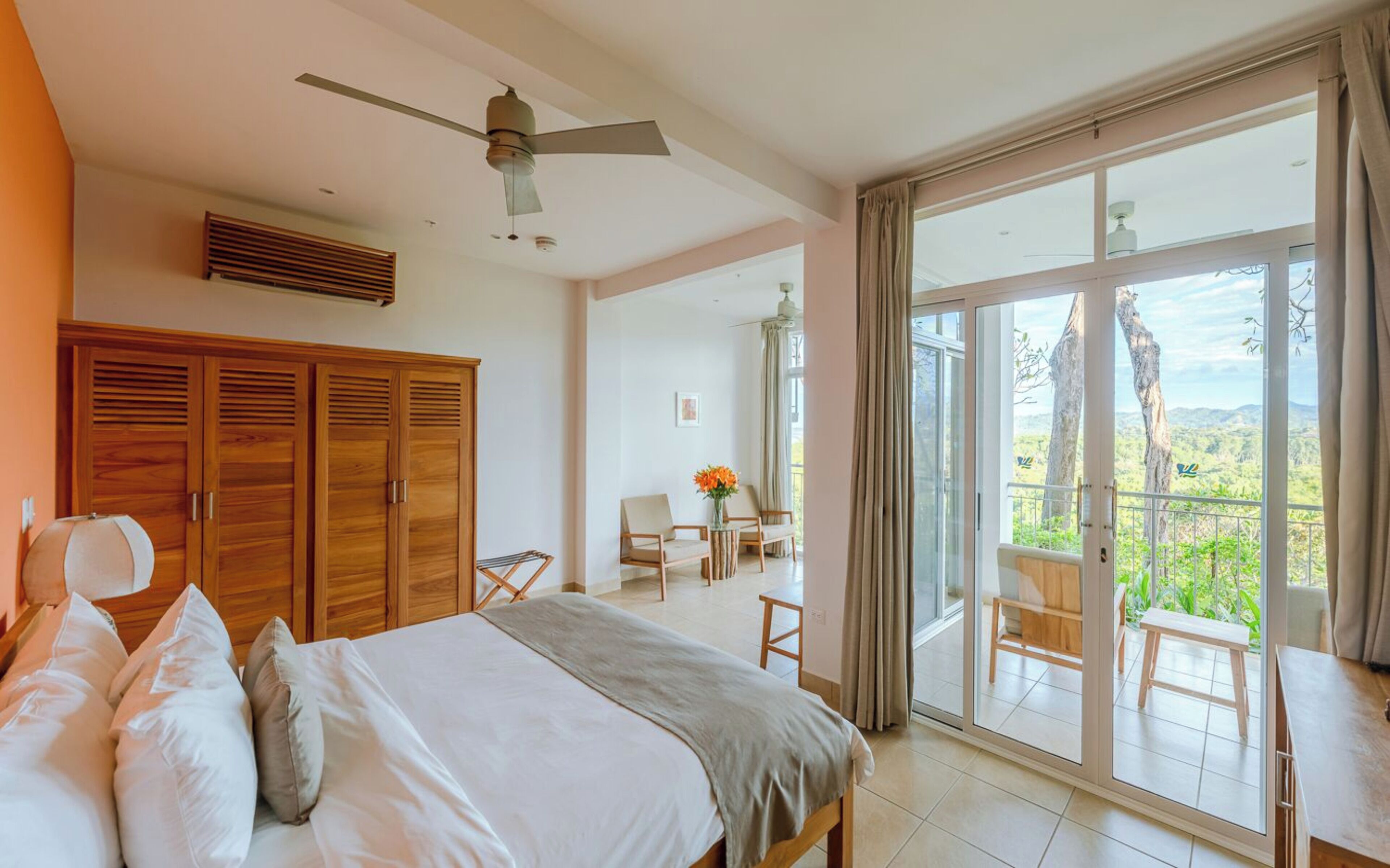 A bedroom with a bed, wooden wardrobe, ceiling fan, and a balcony with a view of trees and hills.