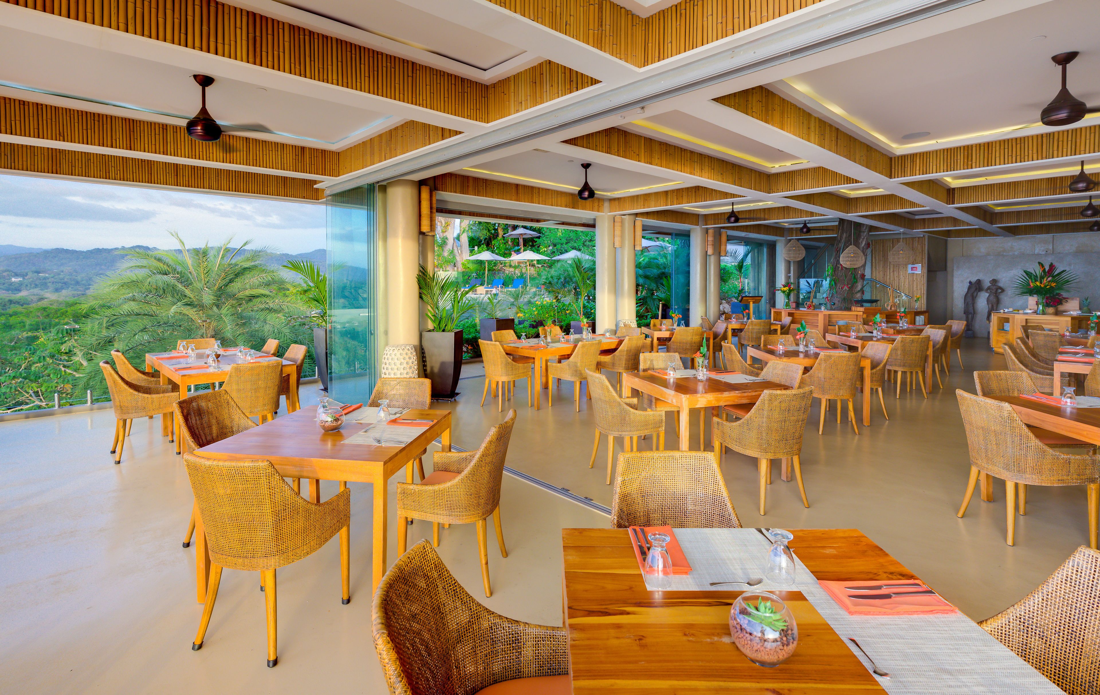 A spacious restaurant with high ceilings, wooden paneling, and large windows offering a view of the outdoors. Tables are set with wicker chairs and dining essentials.