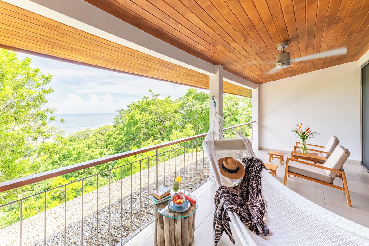 Un balcon avec un hamac, une table avec une boisson et une vue sur l’océan.
