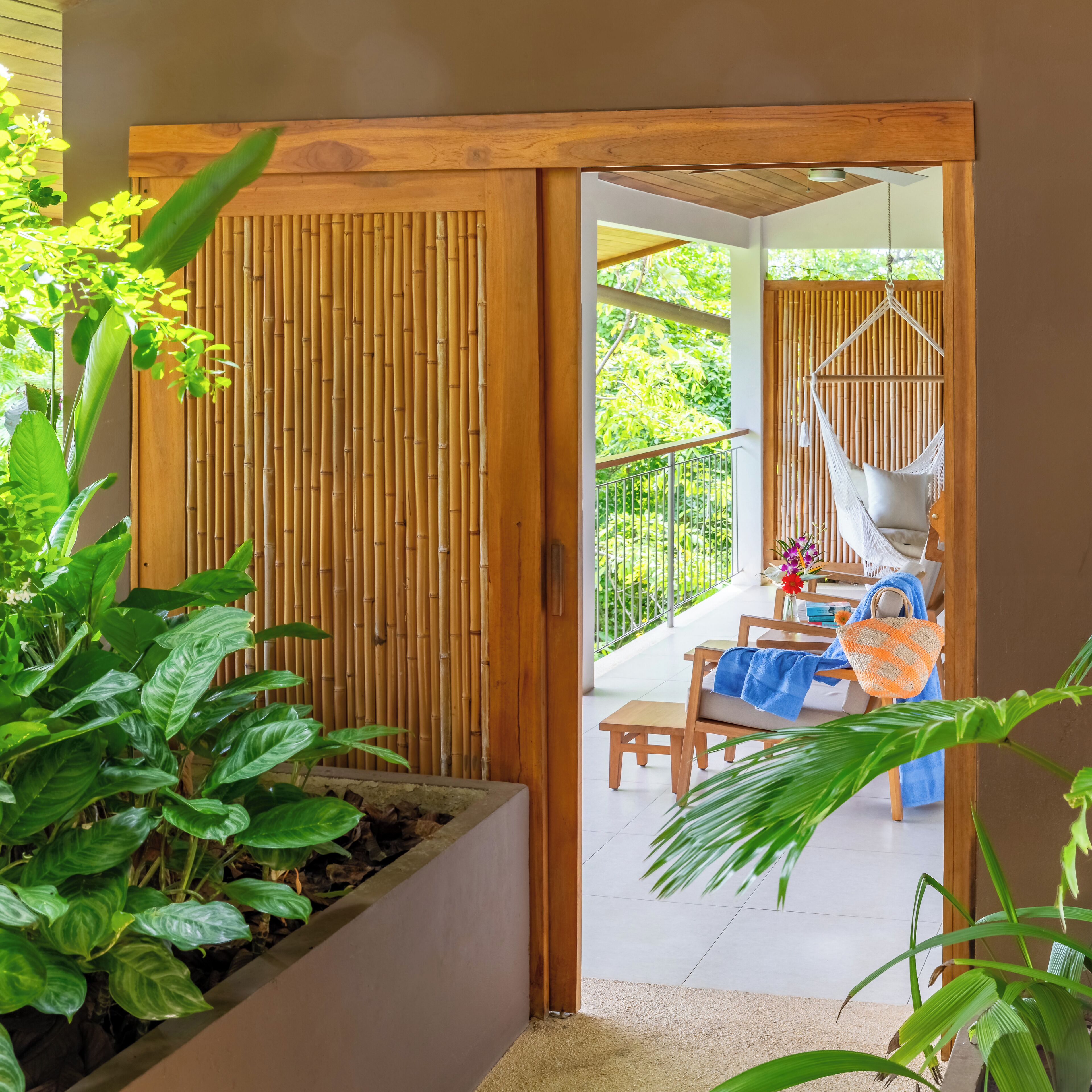A wooden door leading to a balcony with a hammock, a table with a chair, and potted plants.
