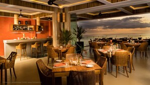 Un restaurante con barra, varias mesas para comer, sillas de mimbre y vista al océano.