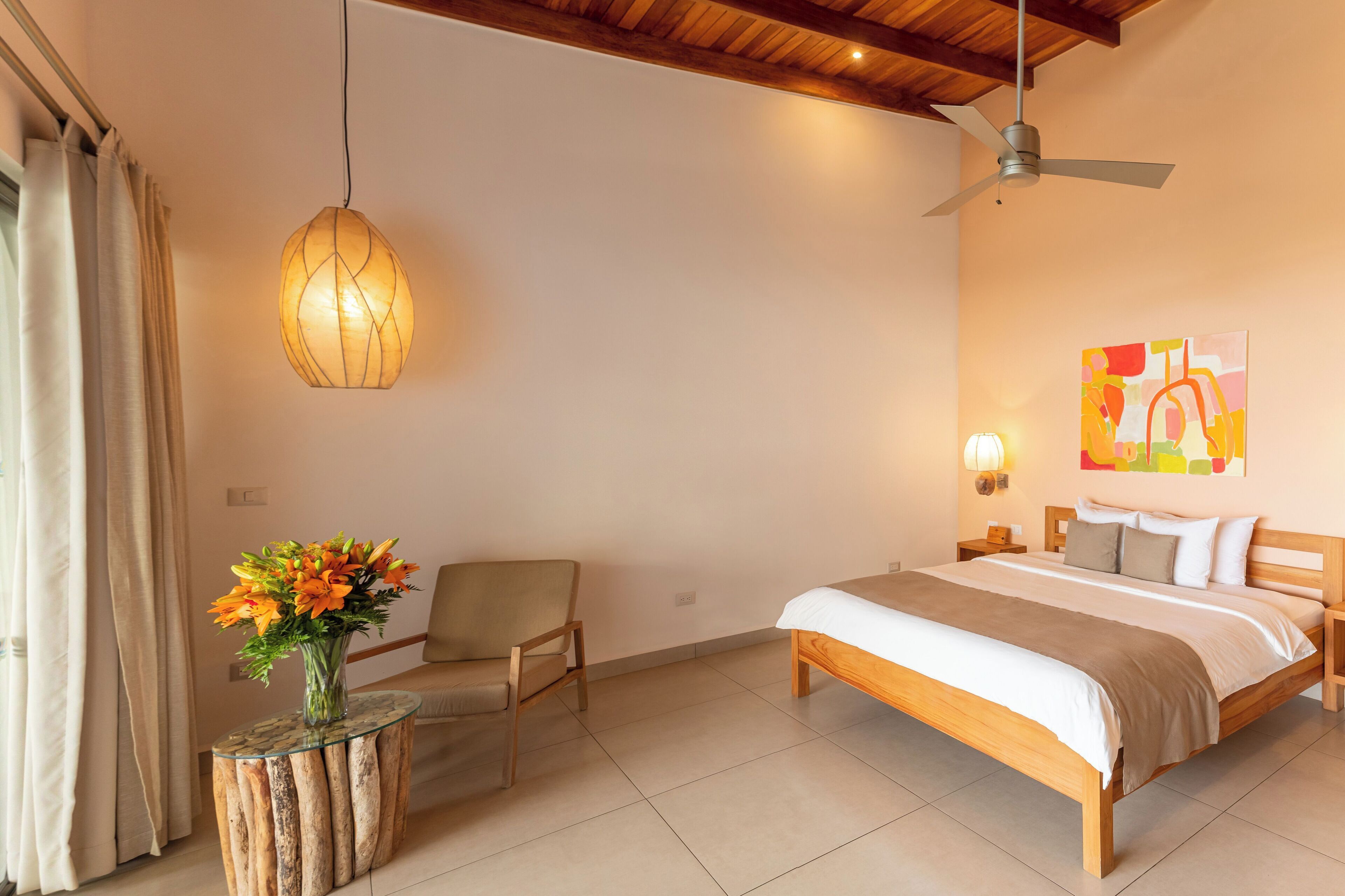 Un dormitorio con cama, una silla, una mesa con un jarrón de flores, un ventilador de techo y una lámpara de pared.