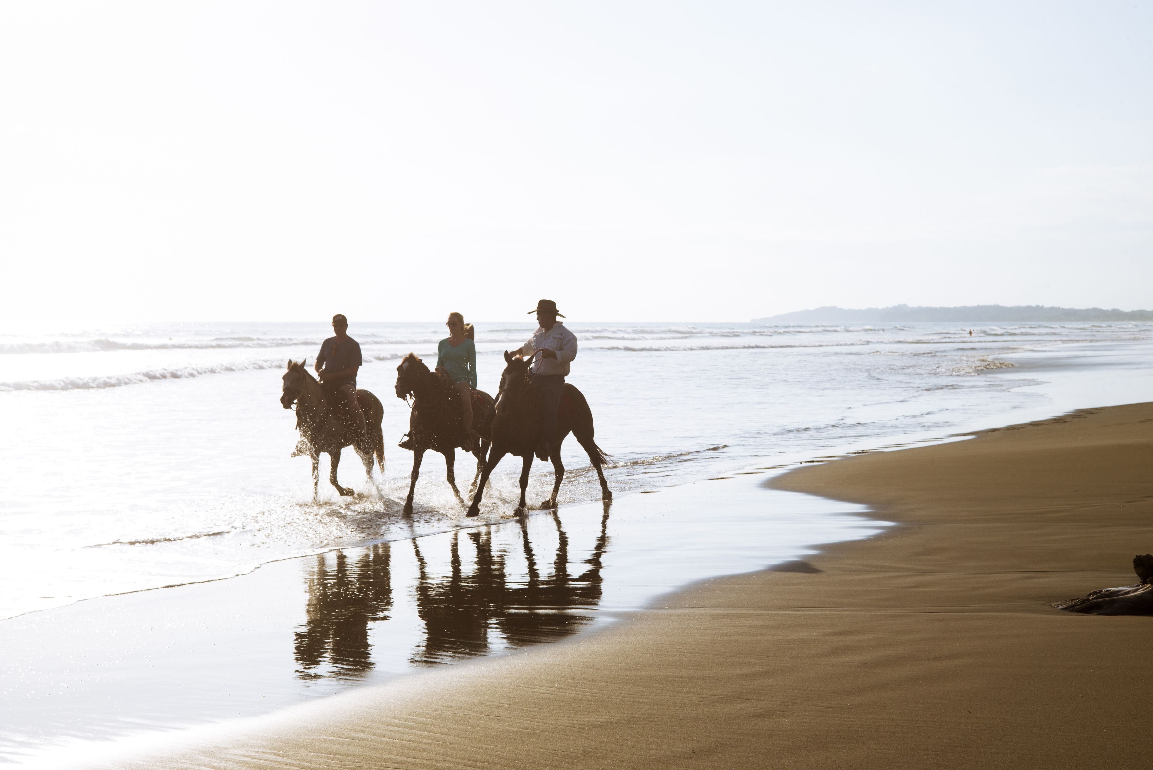 A beach scene with three people riding horses along the shoreline.