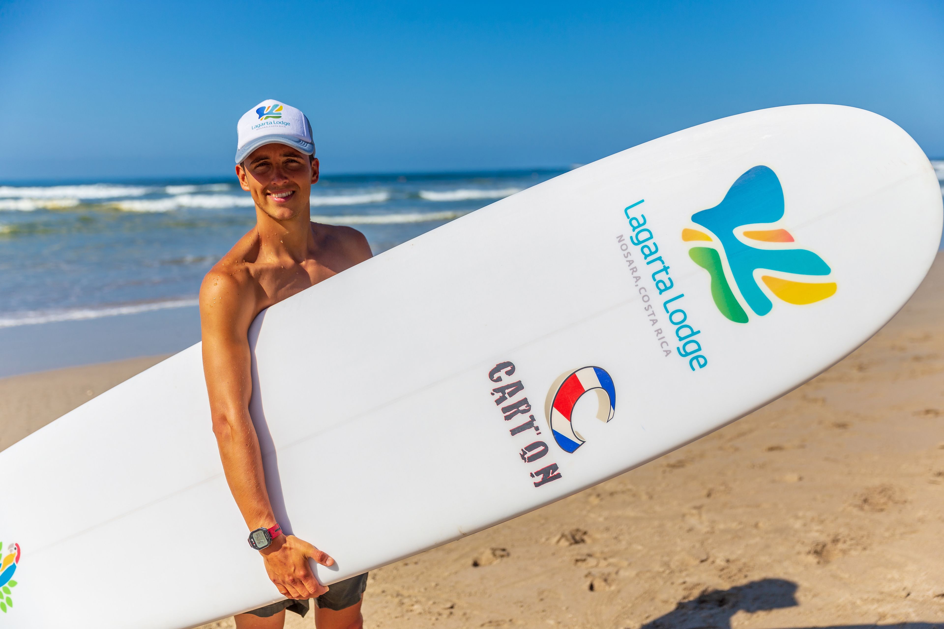 A person holding a surfboard with logos for Laguna Lodge and Carton on a beach.