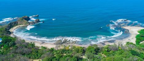 Vista aerea di una zona costiera con spiaggia, affioramenti rocciosi e vegetazione lussureggiante.