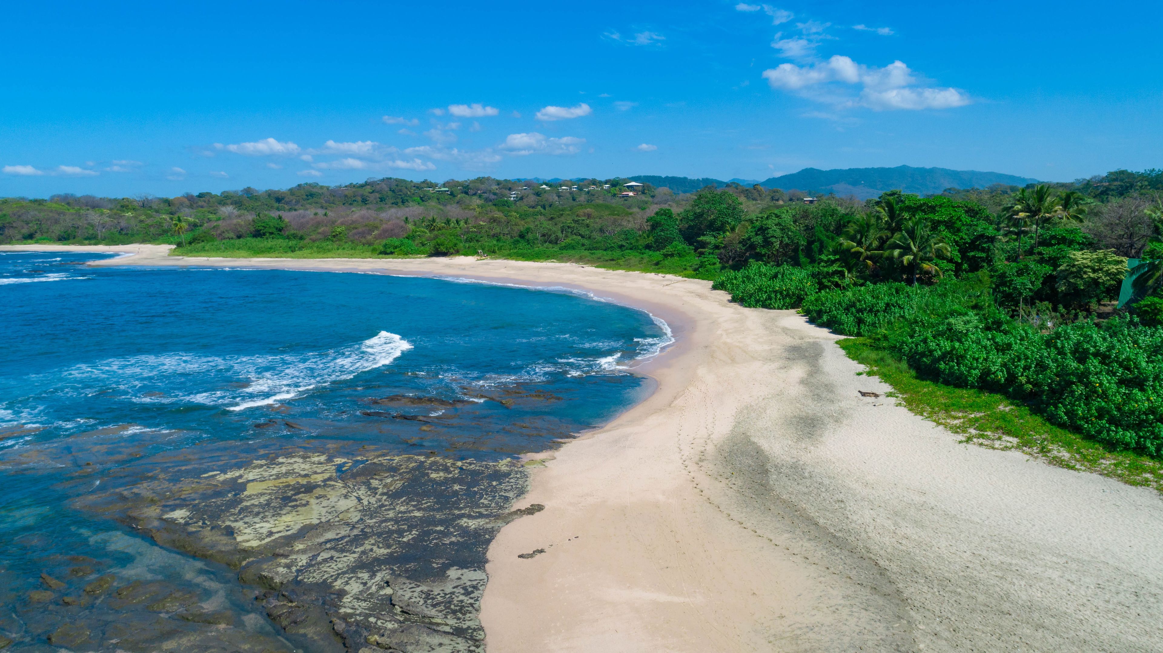 A coastal landscape with a sandy beach, clear blue waters, and lush greenery.