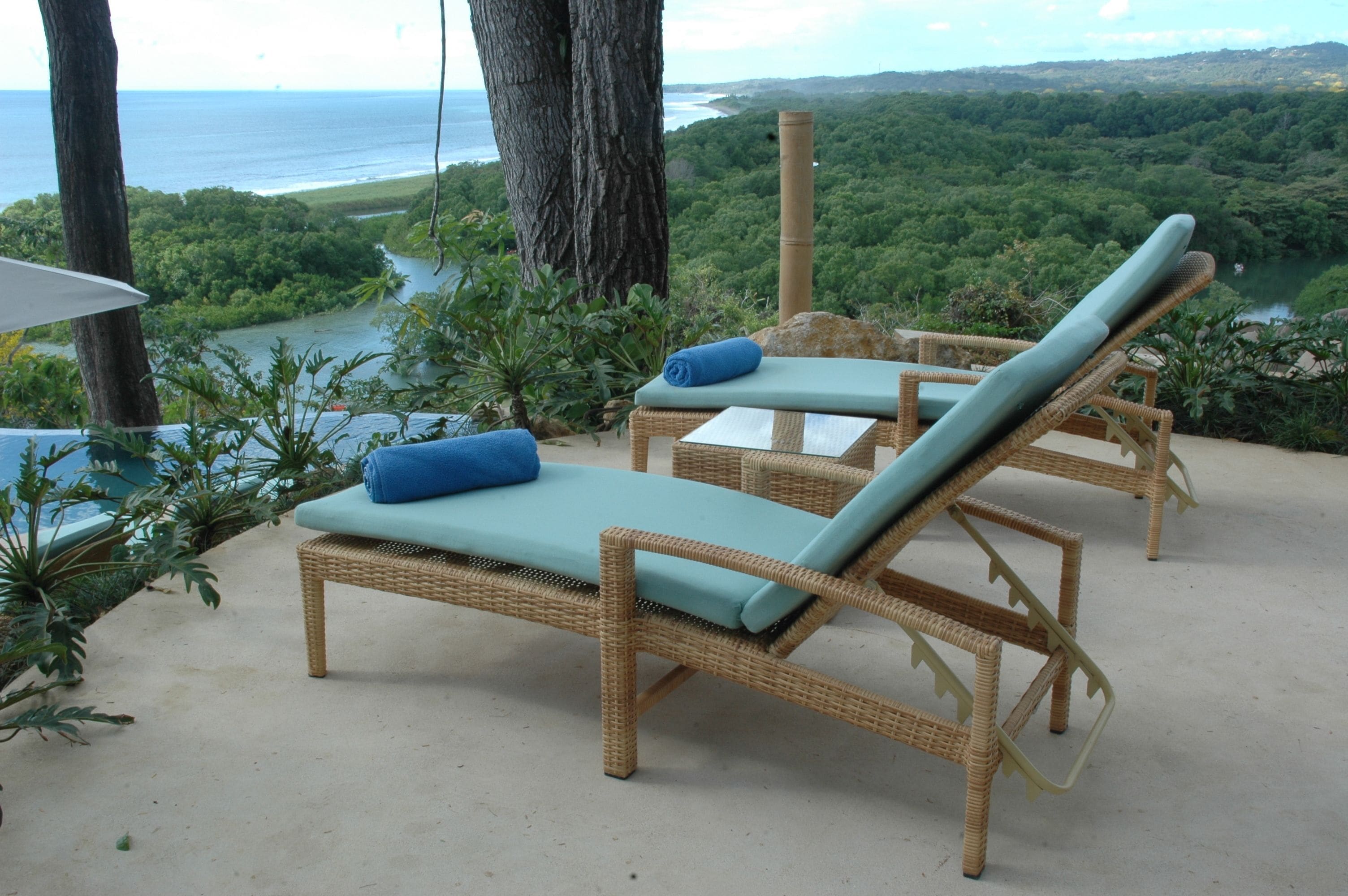 Sillones de mimbre con cojines azules, suelo de hormigón y vistas a un paisaje verde exuberante y el océano.