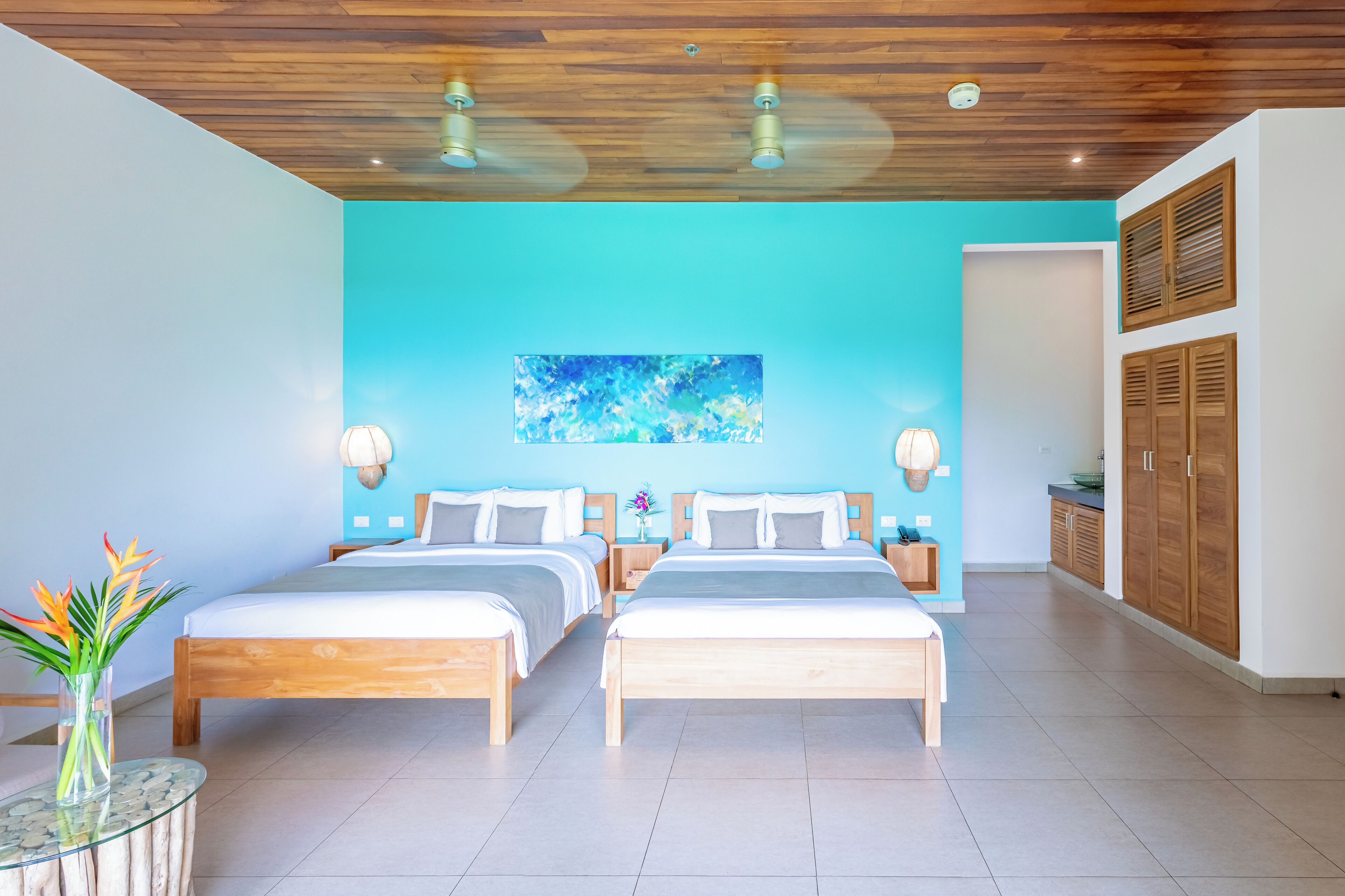 A modern hotel room with two wooden beds, a blue accent wall, and a ceiling fan.