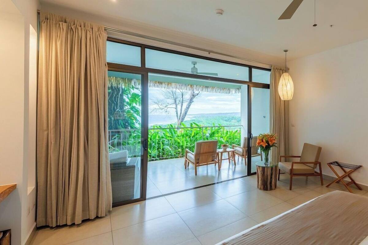 Une chambre d’hôtel moderne dotée d’une grande porte-fenêtre coulissante donnant sur un balcon aménagé avec du mobilier d’extérieur et offrant une vue sur la verdure.