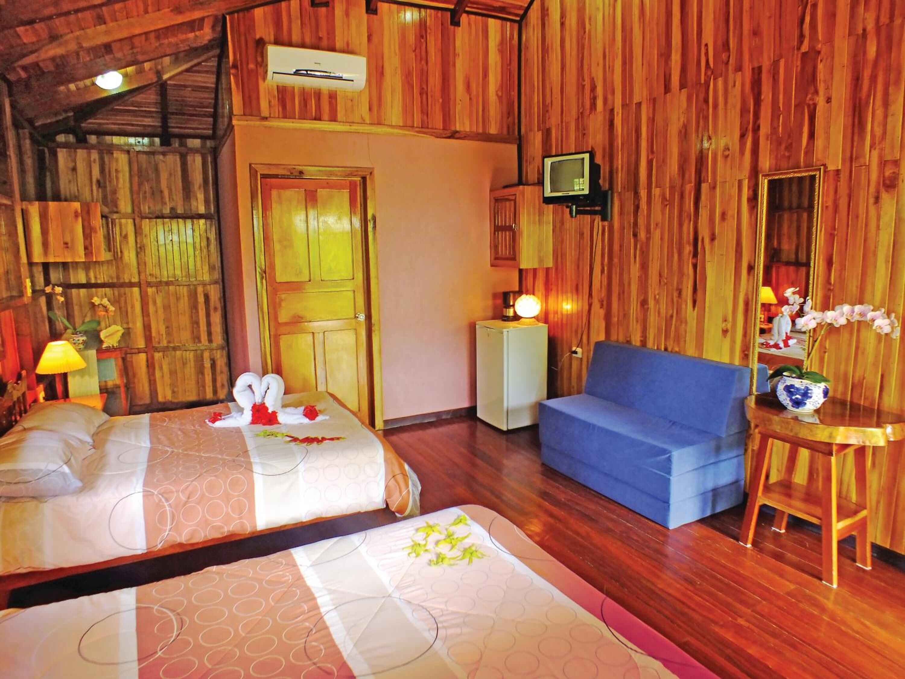 A room with wooden walls and floors, two beds with floral arrangements, a blue sofa, a small table with a lamp, and a television on the wall.