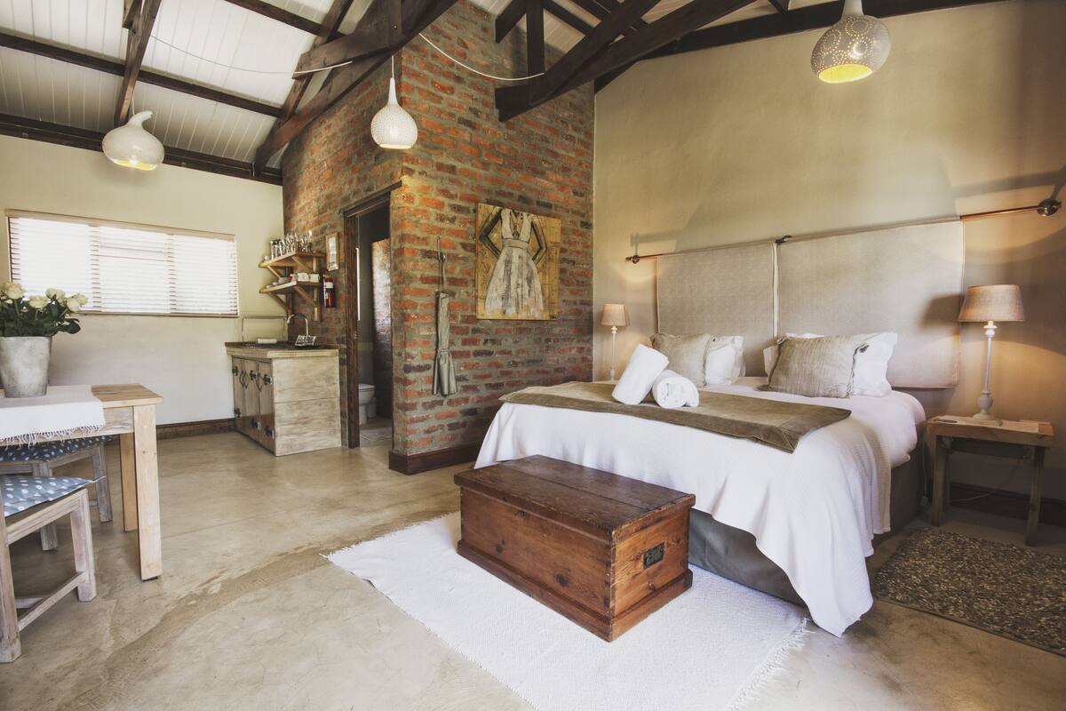 Een eenvoudige slaapkamer met een bakstenen muur, een houten kist, een bed met witte lakens en een hanglamp.