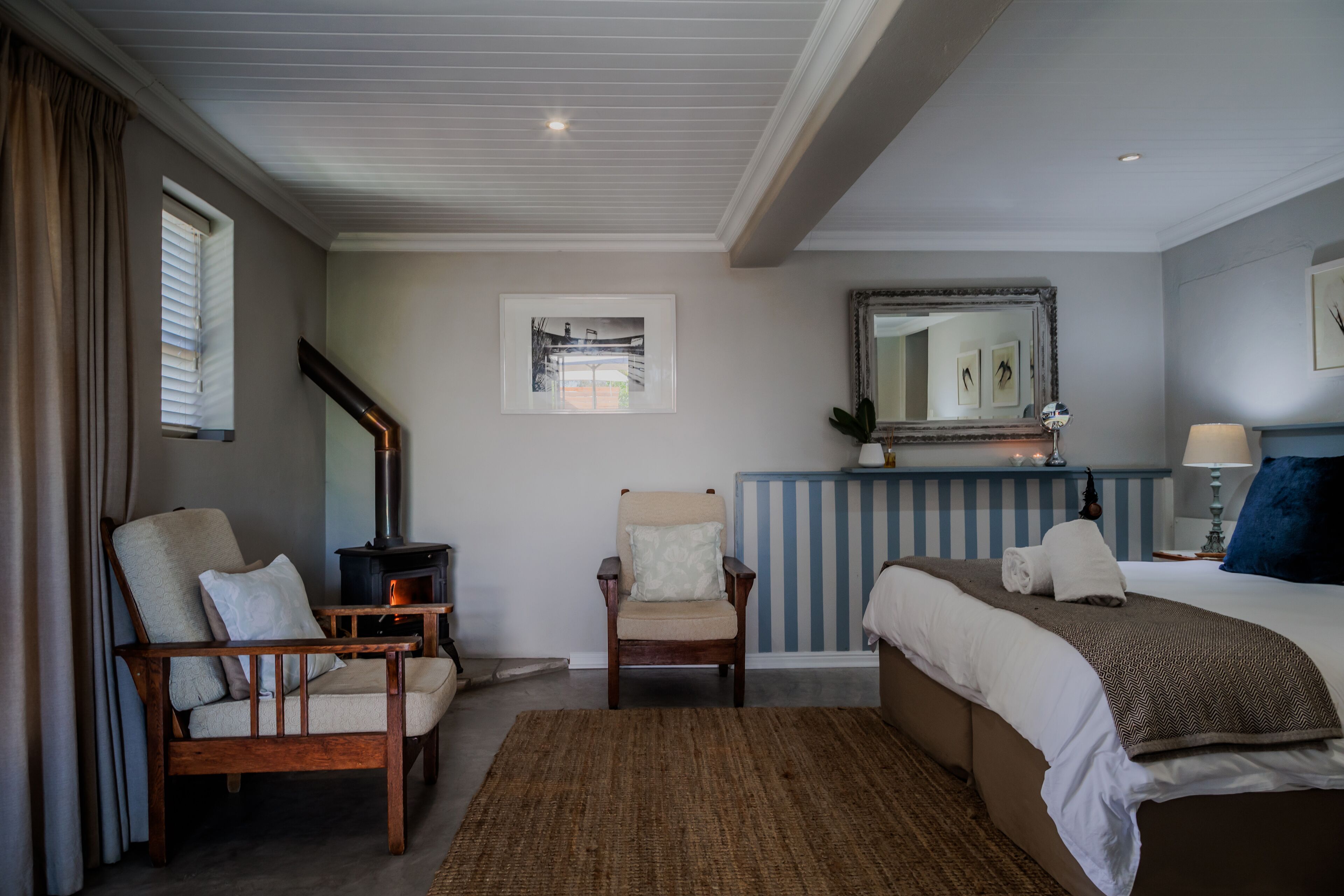 A bedroom with a bed, two chairs, a fireplace, a mirror, and artwork on the wall.