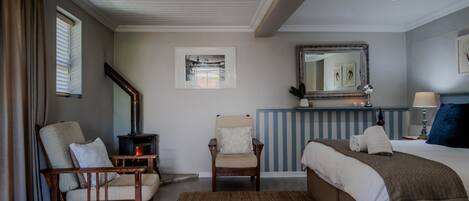 A bedroom with a bed, two chairs, a fireplace, a mirror, and artwork on the wall.