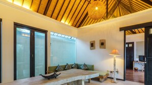 A spacious room with a wooden table, a green sofa with patterned cushions, a hanging light fixture, and a view through a glass door. - Artemis Villa (Seminyak)