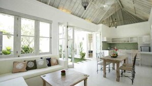 A bright, open-plan living area with a kitchen, large windows, and a ceiling with exposed beams. - Artemis Villa (Seminyak)