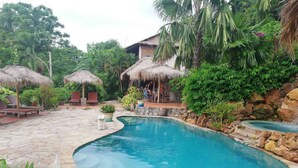A pool area with a curved design, surrounded by stone flooring and lush greenery. There are thatched-roof structures and outdoor seating. - The Beach House (Kep)