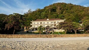 A large, multi-story building with a symmetrical facade, a central entrance, and a surrounding landscaped area with trees and a stone wall. - The Beach House (Kep)