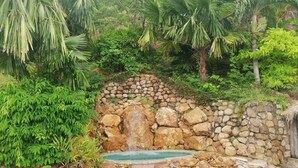 A garden with a stone wall, a waterfall, and lush greenery. - The Beach House (Kep)