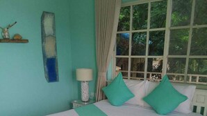 A bed with white bedding and teal accents, a nightstand with a lamp, and a window with curtains. - The Beach House (Kep)