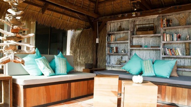 A cozy seating area with a thatched roof, a bookshelf, and a decorative wooden structure. - The Beach House (Kep)