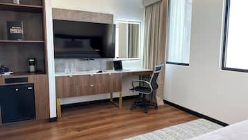 Habitación de hotel moderna con escritorio de madera, televisor de pantalla plana, refrigerador pequeño, cama y ventana grande con cortinas.