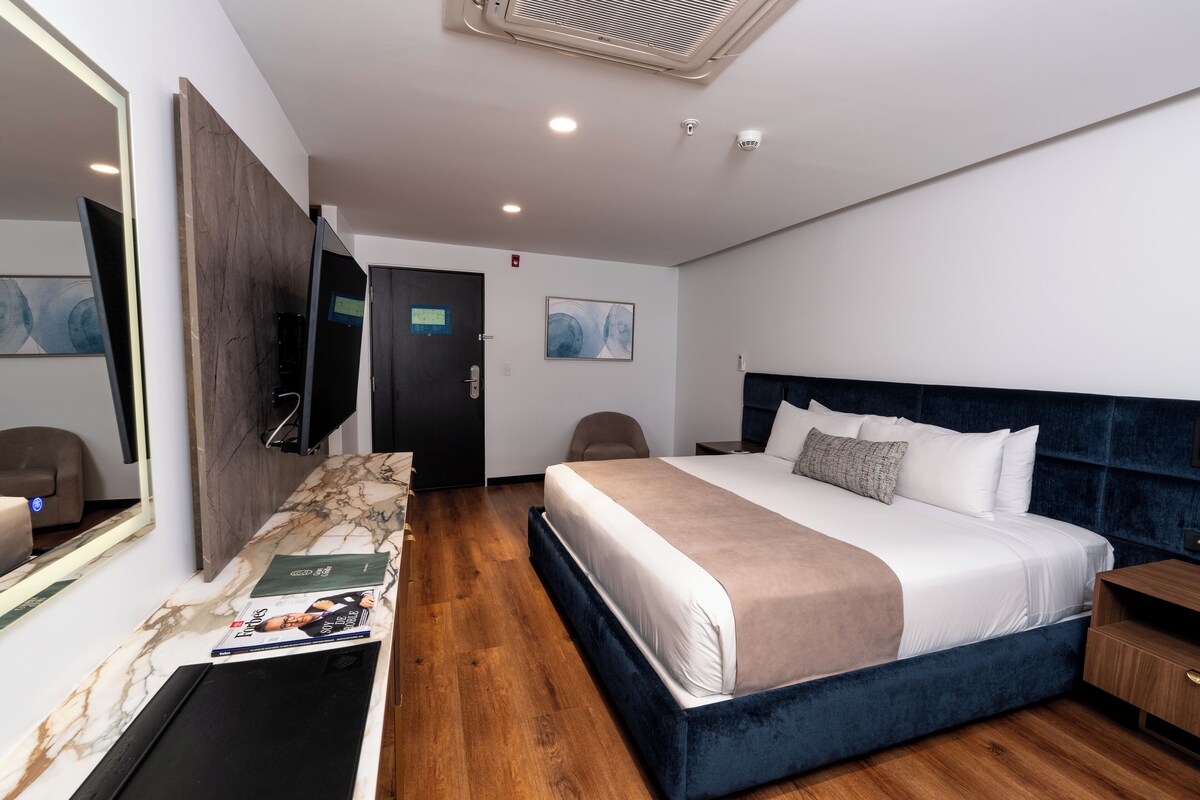 Habitación de hotel moderna con una cama grande, un televisor de pantalla plana, piso de madera y un banco con diseño de mármol.