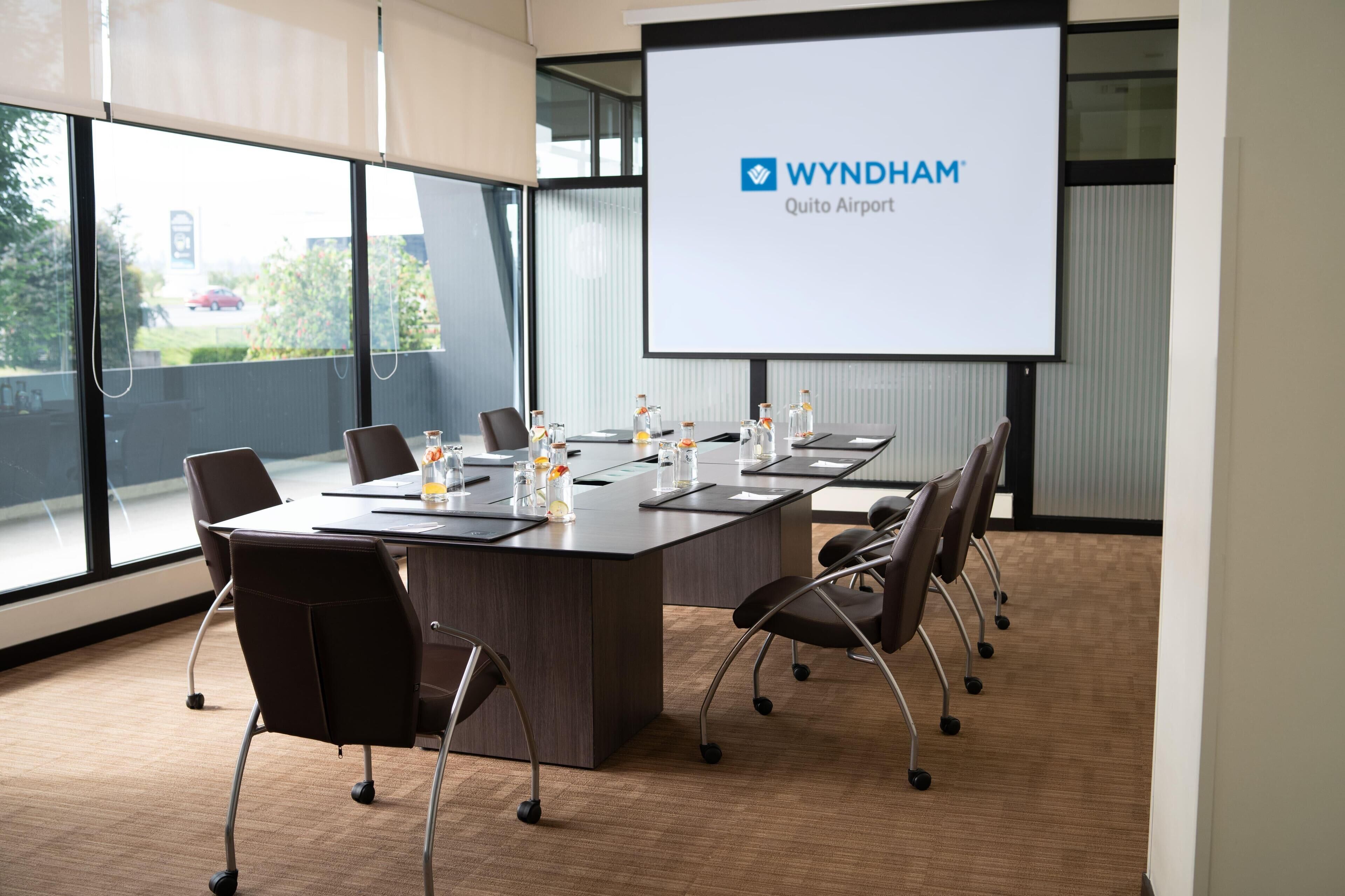 A conference room with a long table, chairs, a projector screen displaying 'WYNDHAM Quito Airport', and a view of the outside.