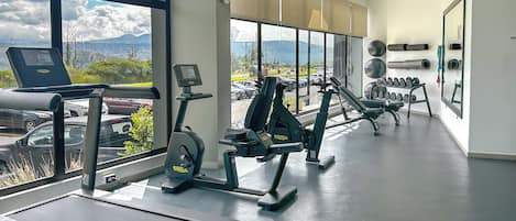 Un gimnasio con equipo de cardio, pesas y un amplio ventanal con vistas a montañas y coches.