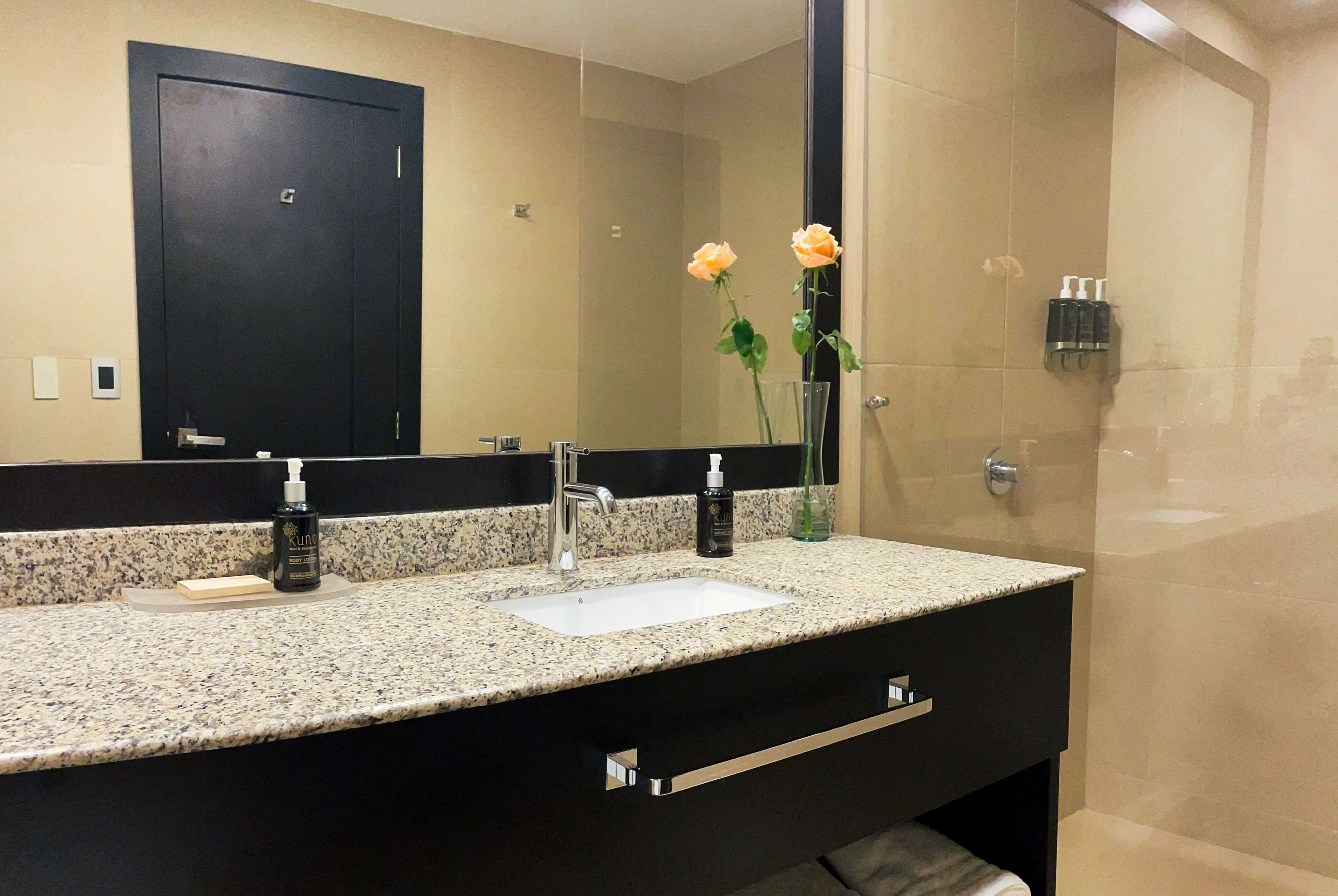 Un baño con encimera de granito, lavabo blanco, mueble negro, espejo y zona de ducha.