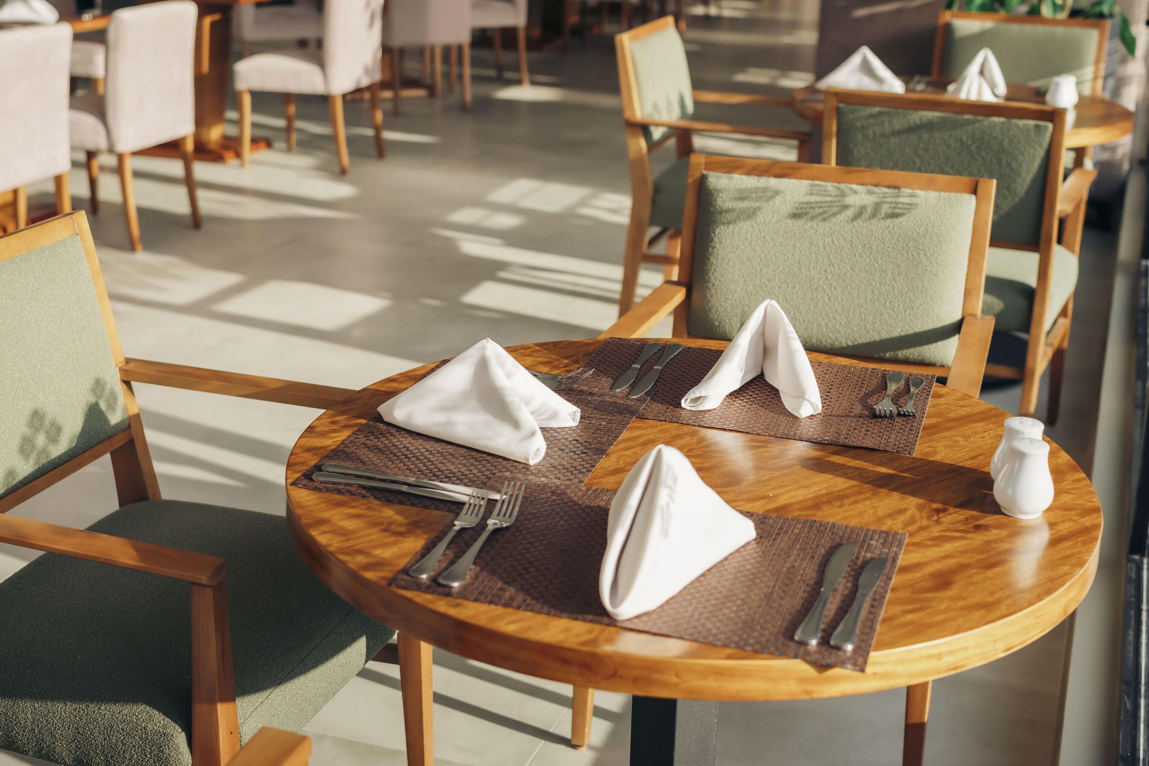 Un comedor con mesas redondas de madera, sillas con cojines verdes y una vajilla ordenada que incluye cubiertos y servilletas.