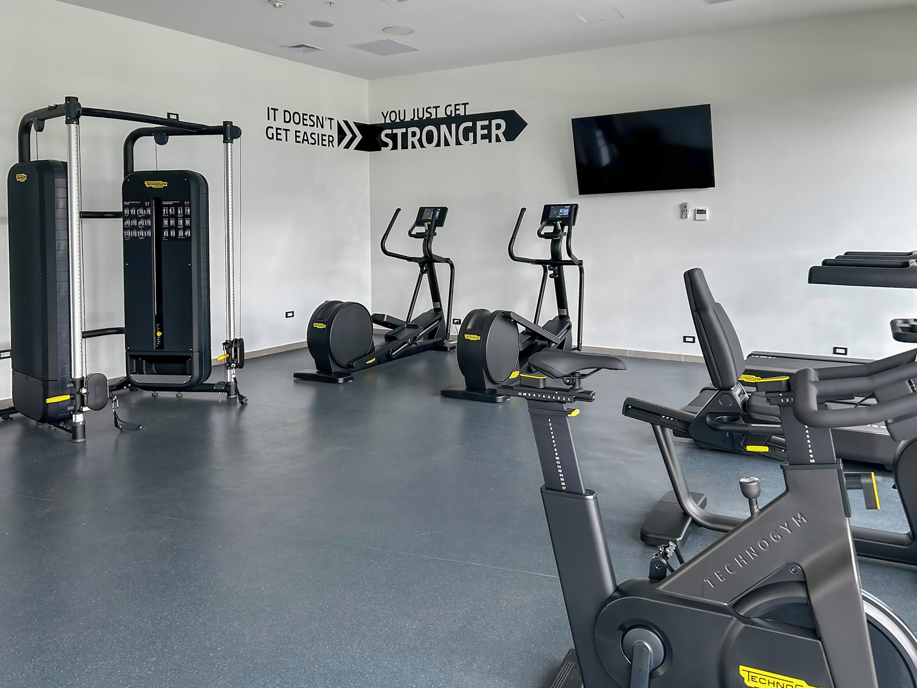 Un gimnasio con equipo de ejercicio, un letrero motivacional en la pared y un televisor montado.