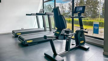 A gym with treadmills, a weight bench, and a rowing machine, with a view of trees and a clear sky through large windows.