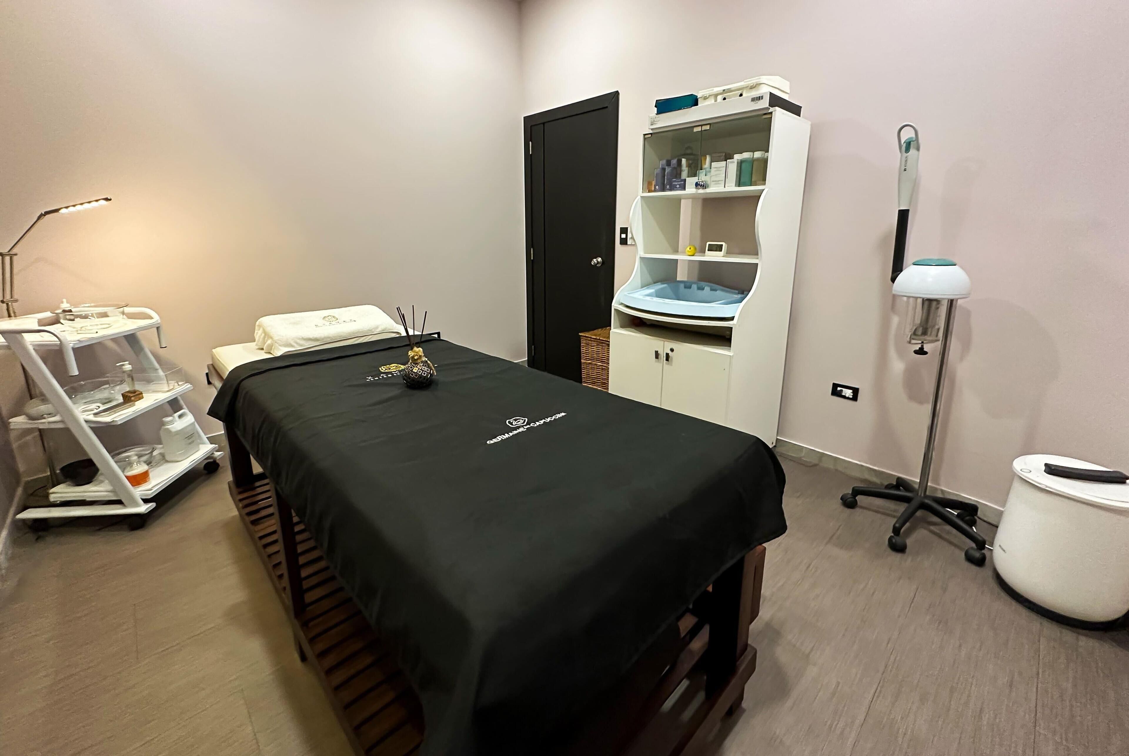 A medical examination room with a treatment bed, a cart with medical supplies, a cabinet with shelves, and a footrest.