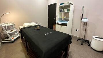 A medical examination room with a treatment bed, a cart with medical supplies, a cabinet with shelves, and a footrest.