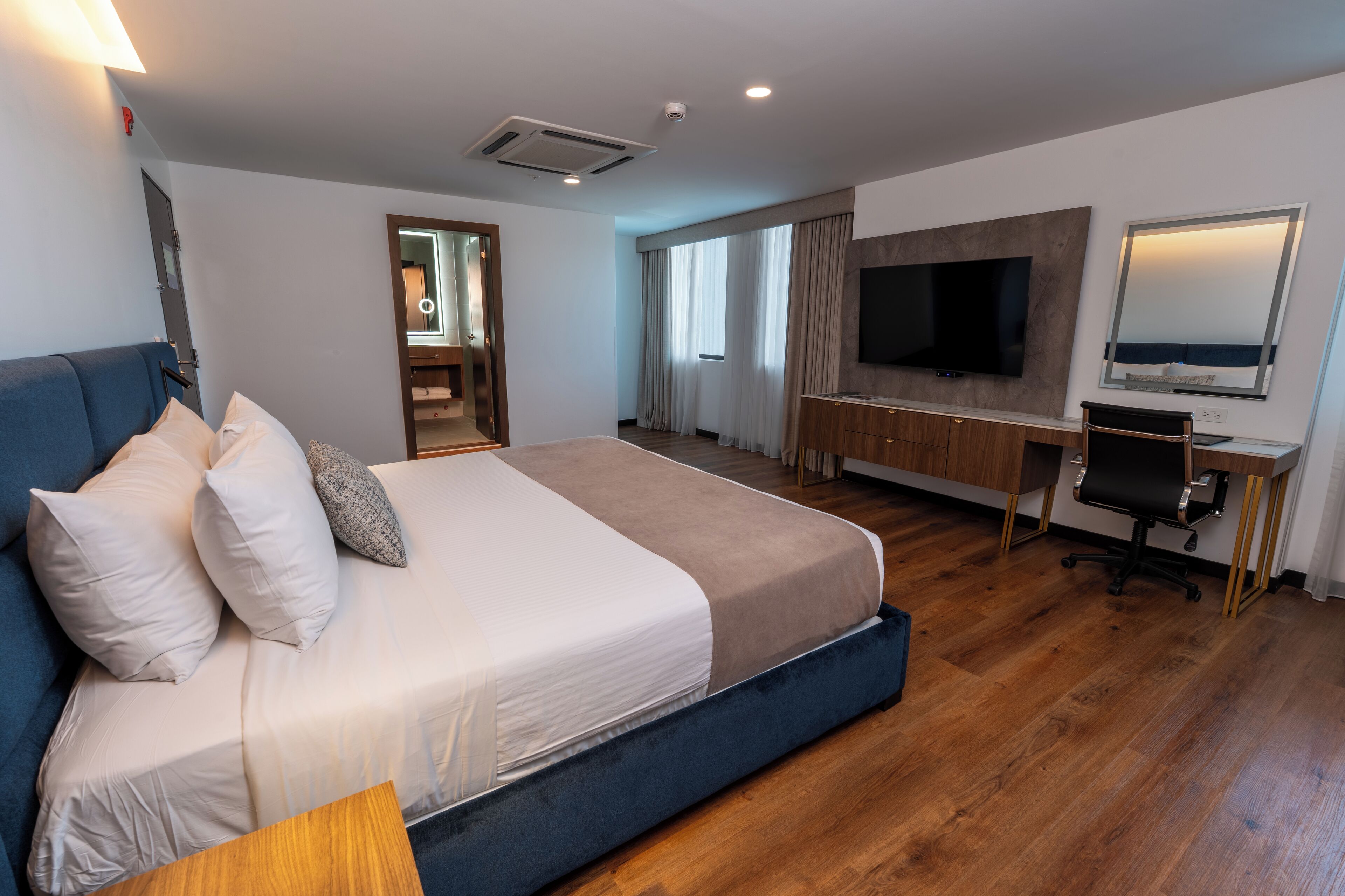 A modern hotel room with a large bed, a desk with a chair, a TV, and a mirror.