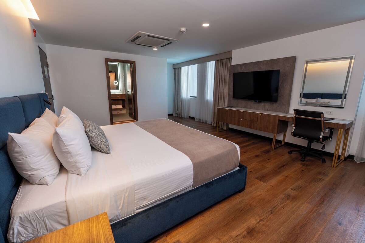 Una habitación de hotel moderna con una cama grande, un escritorio con silla, un televisor y un espejo.