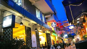 A street view at night with a restaurant on the left, a shop with umbrellas on the right, and a blue sky above. - The Purple Mangosteen (Siem Reap)
