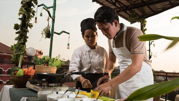 Two chefs preparing food outdoors with a table set for dining, surrounded by greenery and a rustic setting. - The Purple Mangosteen (Siem Reap)