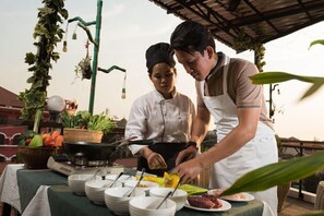 To kokke tilbereder mad udendørs ved et spisebord, omgivet af frodig vegetation og et rustikt miljø.