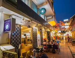 A bustling street scene with outdoor seating at a restaurant, a visible sign for 'The Purple Mangosteen Restaurant & Rooftop', and a variety of shops and stalls.