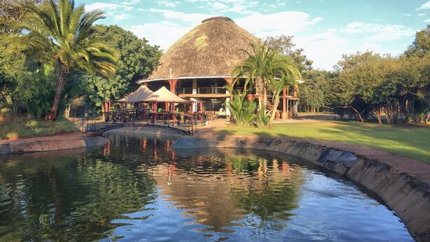 A resort with a thatched-roof building, a swimming pool, and lush greenery. - Kaazmein Lodge (Livingstone)