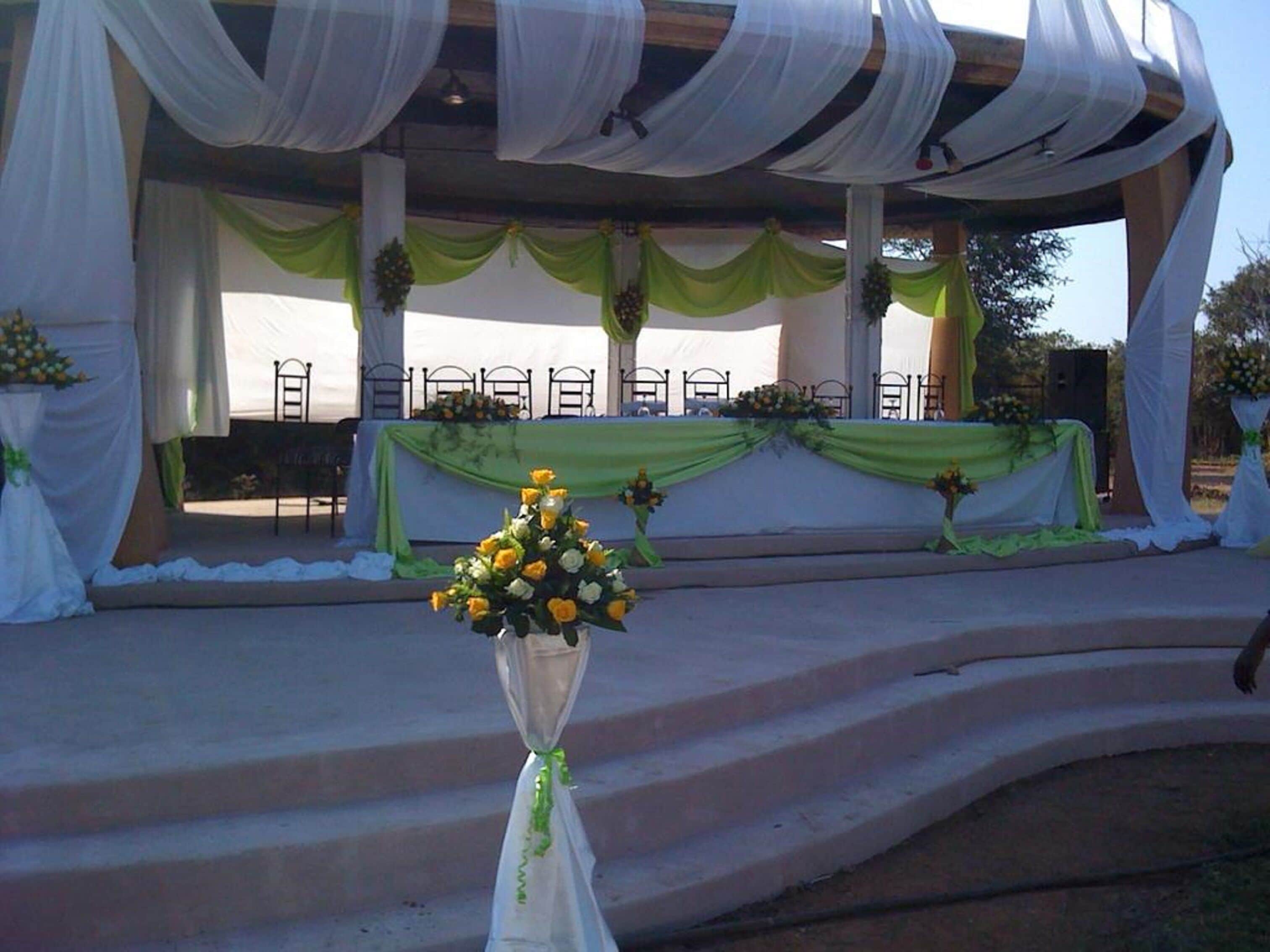 Un escenario al aire libre con cortinas verdes y tela blanca, un arreglo floral en primer plano y equipo de sonido al fondo.