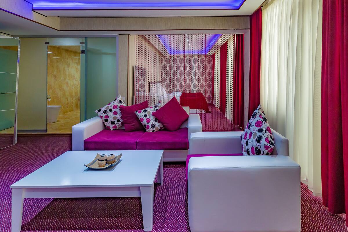 A modern hotel room with a white sofa, a white armchair, a coffee table, and a patterned curtain.