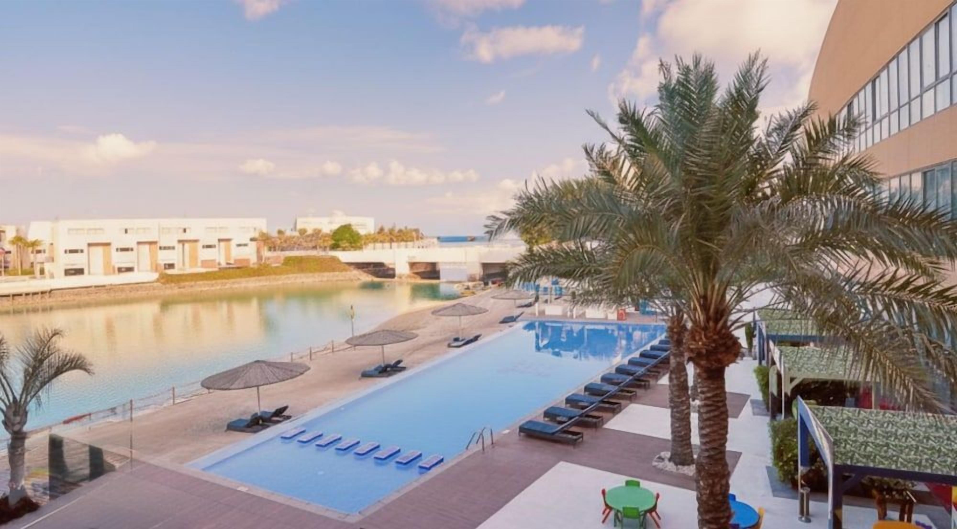 A swimming pool with lounge chairs and umbrellas, a palm tree, and a building with large windows.
