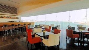 A modern bar area with high stools and a long counter, a dining area with tables and chairs, and a large window overlooking the sea. - The Dragon Beach Hotel & Resort (Amwaj Islands)