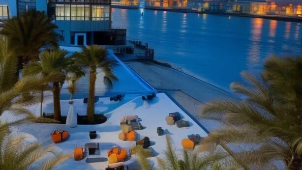 A waterfront hotel with a poolside area, palm trees, and a city skyline in the background. - The Dragon Beach Hotel & Resort (Amwaj Islands)