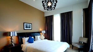 Ein Hotelzimmer mit einem großen Bett, einem Nachttisch, einem Sessel, einer Chandelüster und einem fenster mit Vorhängen.