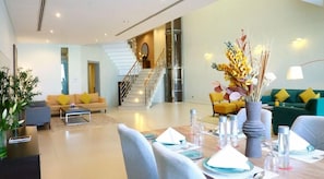 A modern living room with a staircase, yellow and green sofas, a dining area with set tables, and a large vase with flowers. - The Dragon Beach Hotel & Resort (Amwaj Islands)