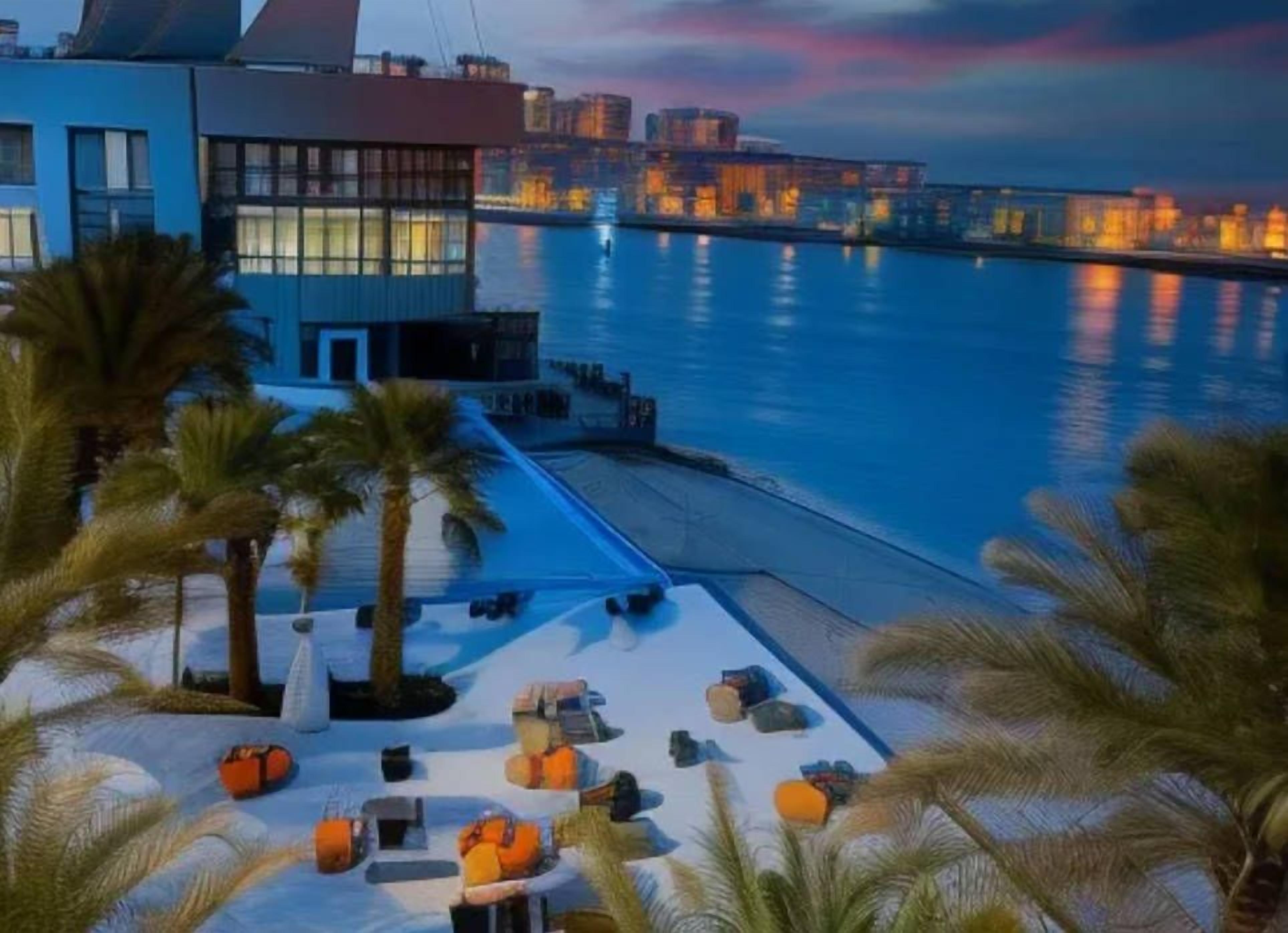 Un hotel frente al mar con zona de piscina, palmeras y el horizonte de la ciudad al fondo.